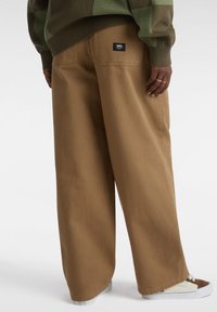 Pantaloni larghi marroni realizzati in tessuto pesante, con una tasca posteriore decorata da un'etichetta nera Vans. Abbinati a sneakers bianche e marroni.