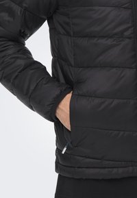 Svart pufferjakke med quiltet design, med en glidelåslomme og elastiske cuffs. Glatt tekstur og lett materiale synlig.