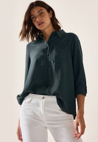 Camisa de botón en azul verdoso oscuro con un bolsillo en el pecho, mangas arremangadas y un ajuste relajado, combinada con pantalones blancos. Tela suave con una ligera textura.