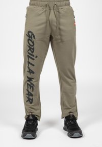 Olijfgroene joggers met een trekkoord in de taille, voorzien van zwarte tekst "GORILLA WEAR" langs het linker been en zijzakken. Draagt zwarte sportschoenen.