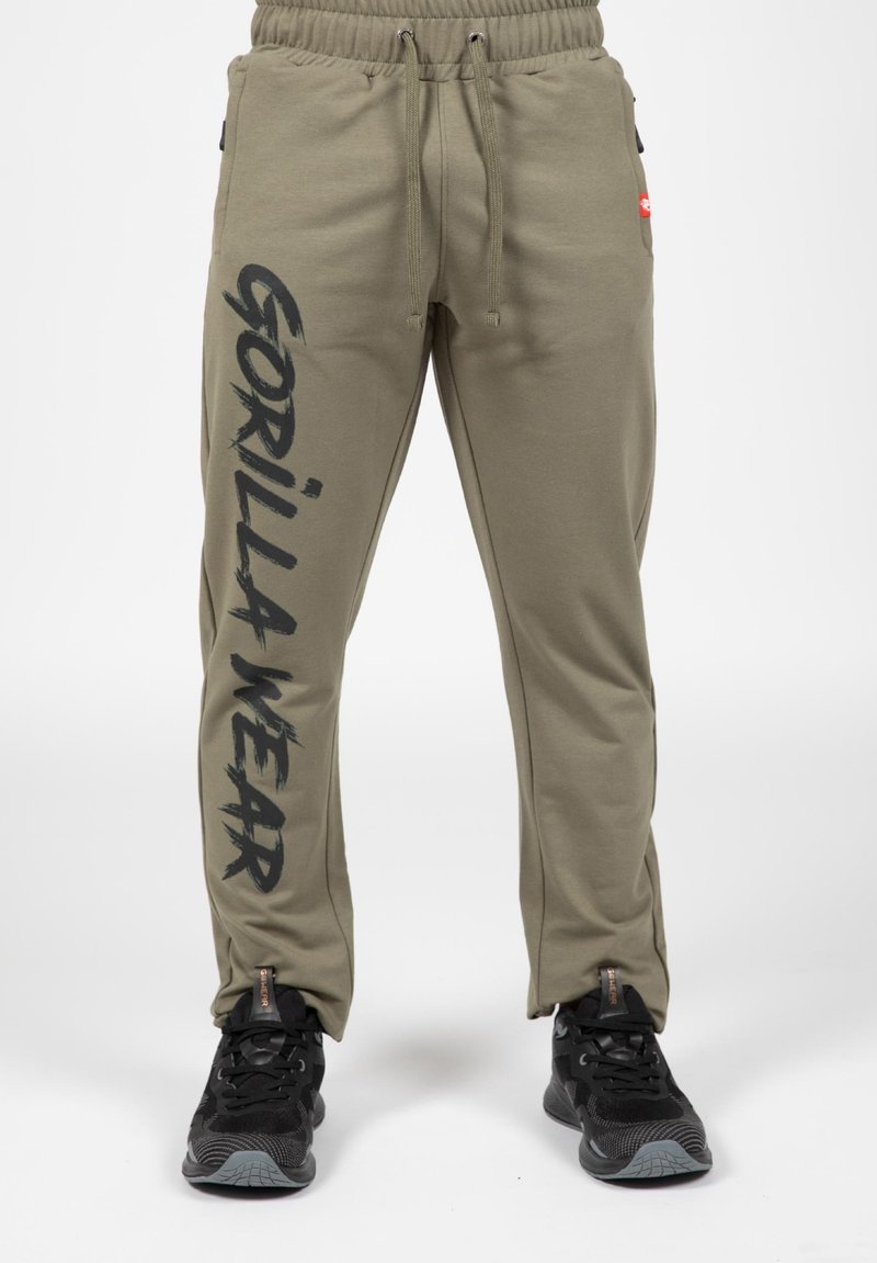 Olijfgroene joggers met een trekkoord in de taille, voorzien van zwarte tekst "GORILLA WEAR" langs het linker been en zijzakken. Draagt zwarte sportschoenen.