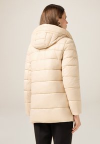 Chaqueta beige acolchada con capucha, que presenta rayas acolchadas, mangas largas y un corte recto, mostrada desde atrás sin cierres visibles.