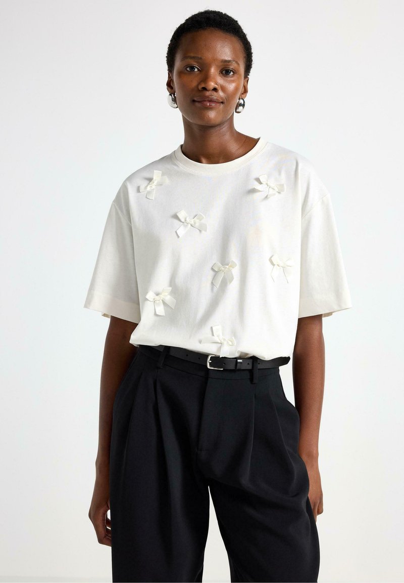 T-shirt bianco a maniche corte con fiocchi in stoffa applicati sul fronte. Abbinato a pantaloni neri a vita alta e una cintura nera.