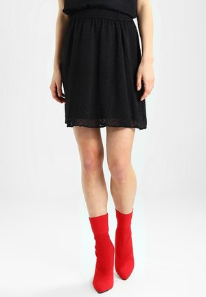 Frau steht, trägt einen schwarzen, strukturierten Rock und leuchtend rote spitze Sockenstiefel vor weißem Hintergrund.