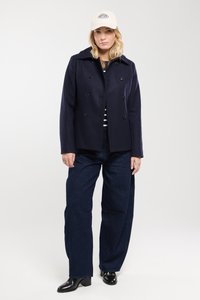 Manteau bleu marine croisé avec des boutons noirs, associé à un haut rayé et un pantalon en denim foncé à jambes larges. Complété par une casquette de baseball crème.