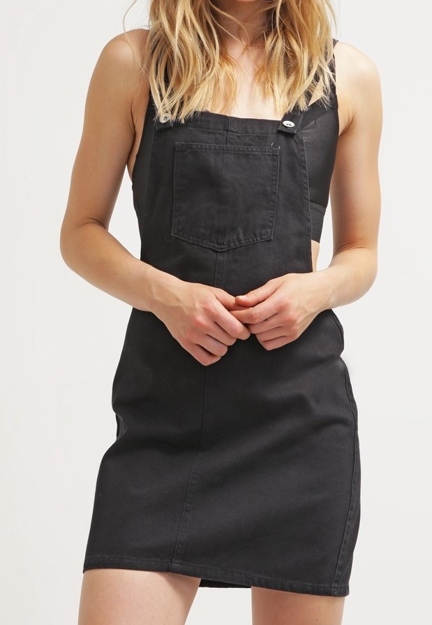 Femme portant une robe salopette en denim noir avec poche avant, debout les mains jointes à la taille devant un fond uni.