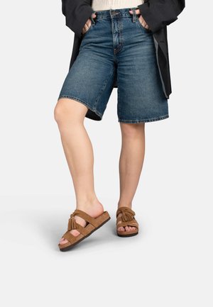 Persona que lleva pantalones cortos de mezclilla azul hasta la rodilla, chaqueta negra y sandalias de ante marrón, de pie con las piernas cruzadas.