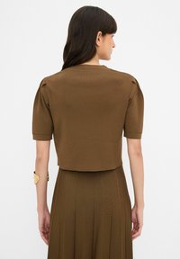 Hnědý žebrovaný pletený crop top s krátkými rukávy a vycpanými ramenními částmi. Zároveň je zadní část hladká a materiál má jemnou texturu.