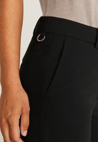 Musta housut, joissa on sileä pinta, klassinen leikkaus, vinoa taskua ja hopeinen rengasvito vyötäröllä.