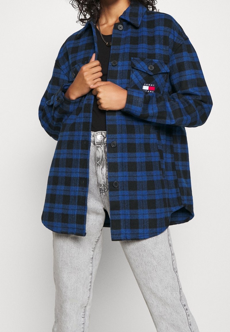 Personne portant une chemise à carreaux bleue et noire boutonnée par-dessus un haut noir avec un jean taille haute gris clair, tenant le devant de la chemise.
