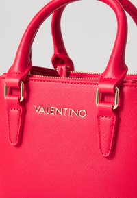 Mazs rozā rokassomu ar strukturētu ārpusi, zelta aksesuāriem un "VALENTINO" zīmolu priekšpusē. Somai ir divi īsi rokturi un rāvējslēdzēja aizdare.