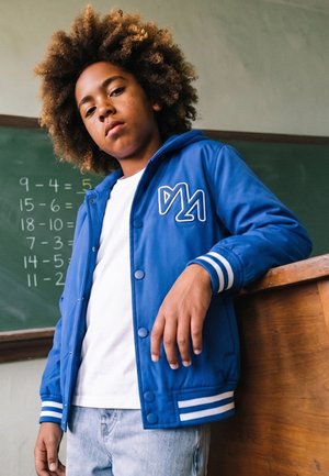 Joven seguro de sí mismo con pelo rizado y chaqueta azul apoyado en un escritorio de madera frente a una pizarra con problemas matemáticos de resta.