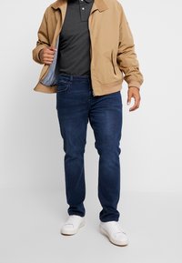 Khaki bomber jacket over a dark grey polo, paired with blue jeans and white sneakers. The jacket features a sturdy collar and zippers.