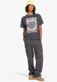 Camiseta oscura gris de corte suelto con estampado gráfico colorido, combinada con pantalones cargo holgados y un gorro liviano verde claro, zapatos tipo slip-on de color marrón claro.