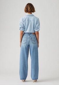 Person with short brown hair wearing a light blue rolled-up long sleeve shirt, high-waisted wide-leg blue jeans, and beige heels, back view.