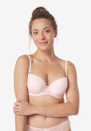 Kobieta z kokiem, ubrana w jasnoróżowy koronkowy biustonosz i dopasowane majtki, delikatnie się uśmiecha, trzymając ręce skrzyżowane na torsie.