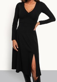 Femme portant une robe noire à manches longues avec col en V et fente frontale, associée à des bottes noires, debout devant un fond uni.