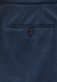 Gros plan sur un tissu bleu marine présentant une poche arrière avec un seul bouton noir et une texture subtile. Détails de couture visibles le long des coutures.