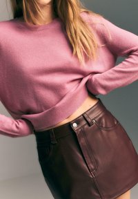 Różowy krótki sweter z okrągłym dekoltem, w zestawieniu z dopasowaną, ciemnobrązową skórzaną mini spódnicą z bocznymi kieszeniami i srebrnymi detalami.