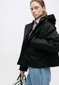 Melns kapuces jaka ar teksturētu флиса oderi, vairākām kabatām un rāvējslēdzēja aizdari. Saskaņota ar gaiši ziliem svītrainiem džinsiem un baltu somu.