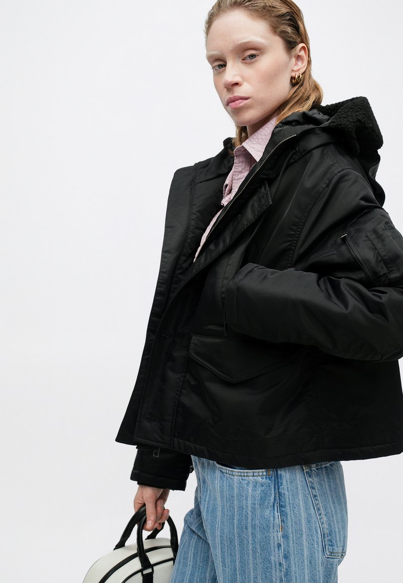 Schwarze Kapuzenjacke mit einem strukturierten Fleecefutter, mehreren Taschen und einem Reißverschluss. Kombiniert mit hellblauen gestreiften Jeans und einer weißen Tasche.