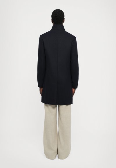 Cămașă din lână bleumarin cu guler înalt, ajustată pe corp, cu mâneci lungi, asortată cu pantaloni largi bej deschis. Textură simplă, design minimalist.