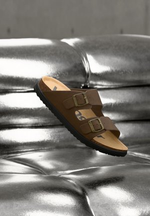 Καφέ σανδάλι με δύο ρυθμιζόμενες αγκράφες και πάτο από φελλό, αιωρούμενο πάνω από μια γυαλιστερή μεταλλική ασημί επιφάνεια με καπιτονάρισμα.