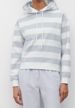 Person trägt einen hellblau-weiß horizontal gestreiften Hoodie mit weißen Kordeln und hellblauen Hosen, steht vor einem weißen Hintergrund.