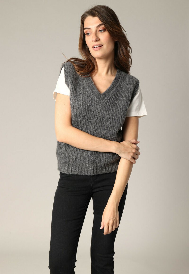 Un gilet sans manches en tricot gris avec un col en V, associé à un t-shirt blanc. Le gilet présente un design ribbed texturé et une silhouette ajustée.