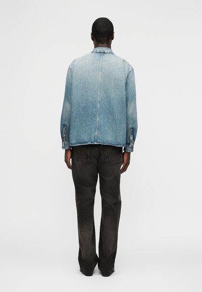 Άνδρας που φοράει ξεθωριασμένο μπλε τζιν μπουφάν και μαύρο τζιν, στέκεται γυρισμένος με την πλάτη προς μια απλή ανοιχτογκρι φόντο.