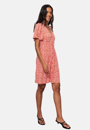 Femme marchant dans une robe rouge fleurie longueur genou avec de courtes manches volantes et des sandales noires à talon bas sur fond blanc.