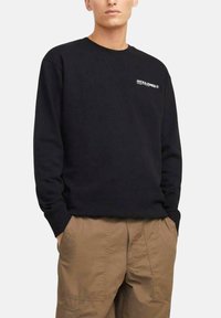 Sudadera negra de manga larga y cuello redondo, con un logotipo blanco en el lado izquierdo del pecho y un corte relajado. Combinada con pantalones cargo beige.