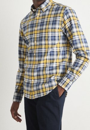 Homme portant une chemise à manches longues à carreaux jaunes, bleus et blancs, un pantalon bleu marine foncé, une main dans la poche, sur un fond uni.