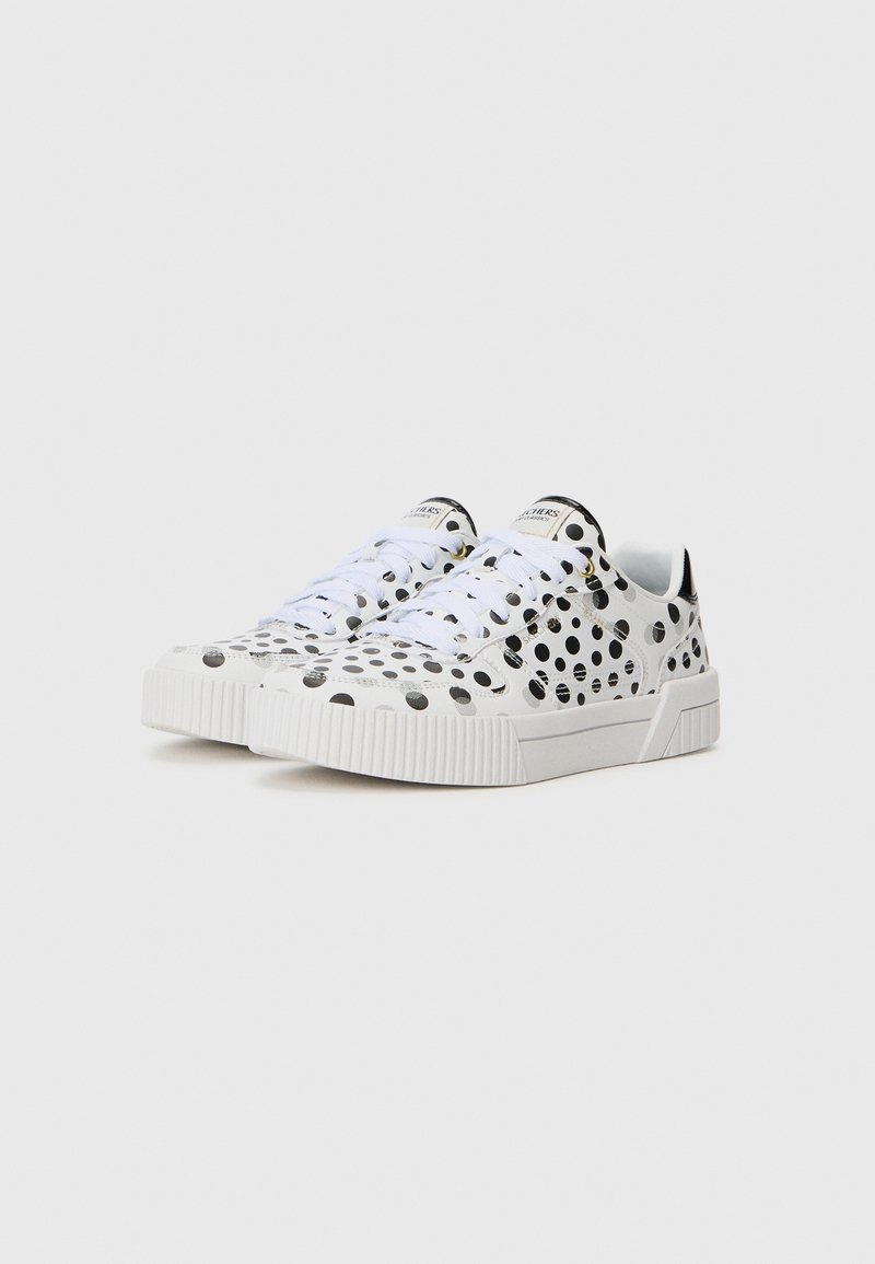 Baskets basses blanches avec des pois noirs, lacets blancs, semelles texturées et étiquette de marque sur la languette et sur le côté.