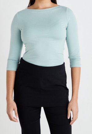 Vrouw met een lichtblauw topje met driekwart mouwen, ingestopt in een zwarte rok over een zwarte broek, tegen een effen witte achtergrond.