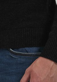 Schwarzer gerippter Strickpullover mit einem enganliegenden Taillenbund, getragen über blauen Jeans. Sichtbare Hand, die sich nahe am Taillenbund ausruht.