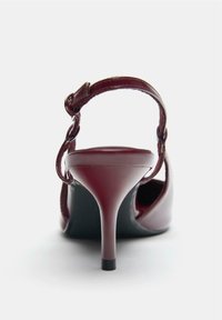 Scarpa con tacco alto in pelle verniciata bordeaux, con cinturino regolabile e mostrata dal retro su sfondo bianco.