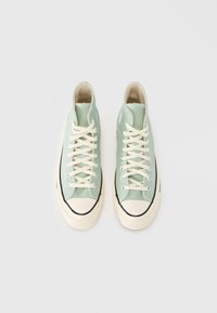 Πράσινα ψηλά πάνινα παπούτσια με μπεζ κορδόνια, λαστιχένιες άκρες δαχτύλων και μαύρες λαστιχένιες σόλες. Διαθέτουν ένα λογότυπο στο εξωτερικό μέρος.