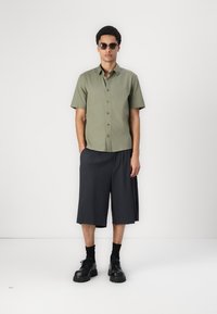 Chemise olive à manches courtes, shorts gris foncé jusqu'aux genoux, chaussures noires chunky et lunettes de soleil ; design sobre avec détails de boutons.