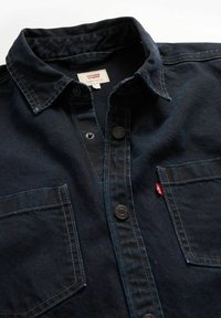 Camisa de mezclilla de color azul oscuro con cuello, dos bolsillos en el pecho y cierres de botones. Presenta costuras en contraste y una pequeña etiqueta de marca.