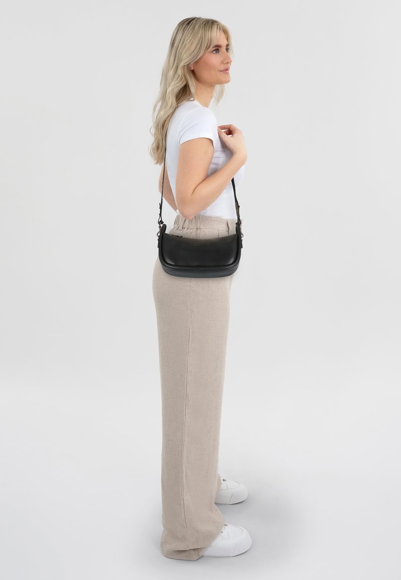 Zwarte leren schoudertas met een gladde afwerking, minimalistisch ontwerp en verstelbare schouderband, gedragen tegen een lichte outfit.