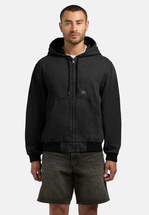 Mann iført en svart hettegenser med glidelås og frontlommer, og mørkegrå denimshorts, stående mot en ensfarget bakgrunn.