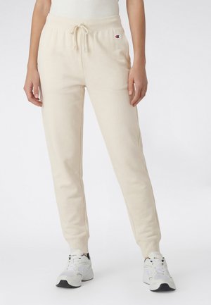 Beżowe spodnie dresowe ze ściągaczem w pasie i elastycznymi mankietami, z logo Champion na lewej udzie, noszone z białymi trampkami na stojącej osobie.