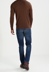 Brun pullovertröja, slim fit, kombinerad med blå denimjeans. Ämnet bär bruna skor, sett bakifrån mot en neutral bakgrund.