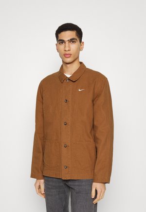 Brown canvas jacket with a collared neck, button closure, two front pockets, and a small white logo on the chest.