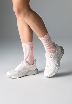 Person trägt weiße und pinke Sportschuhe mit strukturierten Sohlen und pinke Rippsocken mit grauen Unendlichkeitssymbolen.