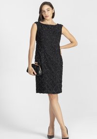 Kobieta stoi z ręką na biodrze, ubrana w bezrękawową czarną sukienkę w kwiatowy wzór do kolan, trzyma czarną kopertówkę i ma na sobie czarne szpilki.