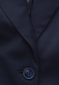 Gros plan sur le revers d'un blazer bleu marine foncé avec un bouton brillant à quatre trous fixé au centre.