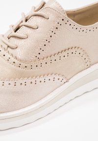 Chaussure brogue en cuir beige clair avec détails perforés, coutures en zigzag, lacets ronds et semelle en caoutchouc blanche.