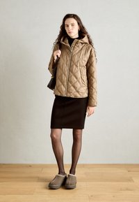Giacca a vento marrone trapuntata con cappuccio, zip argentata e motivo a diamante, abbinata a una gonna nera a matita e a delle scarpe slip-on marroni.